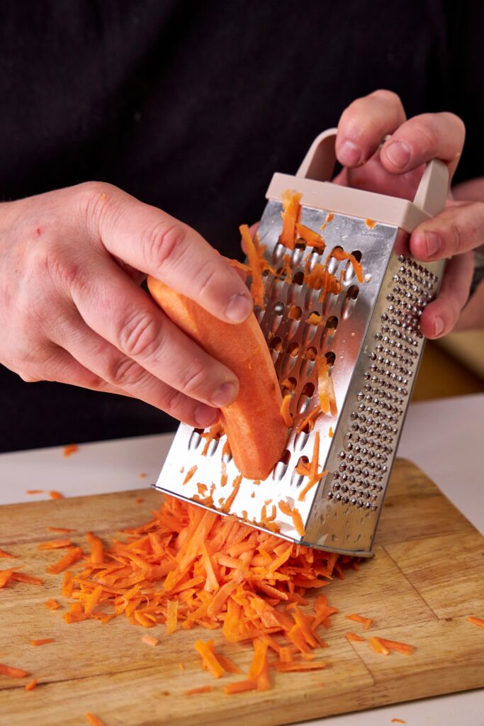 A man in a black shirt grates a carrot using a silver metal box grater