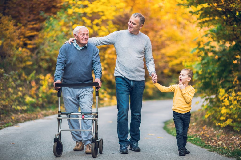 Un père senior marchant avec un dispositif d'aide à la mobilité, un fils adulte et un petit-fils en promenade dans le parc.