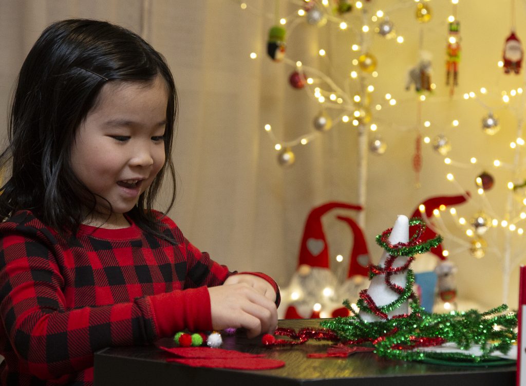 A girl in red plaid sweater playing with a toy at a table with Christmas lights in background.