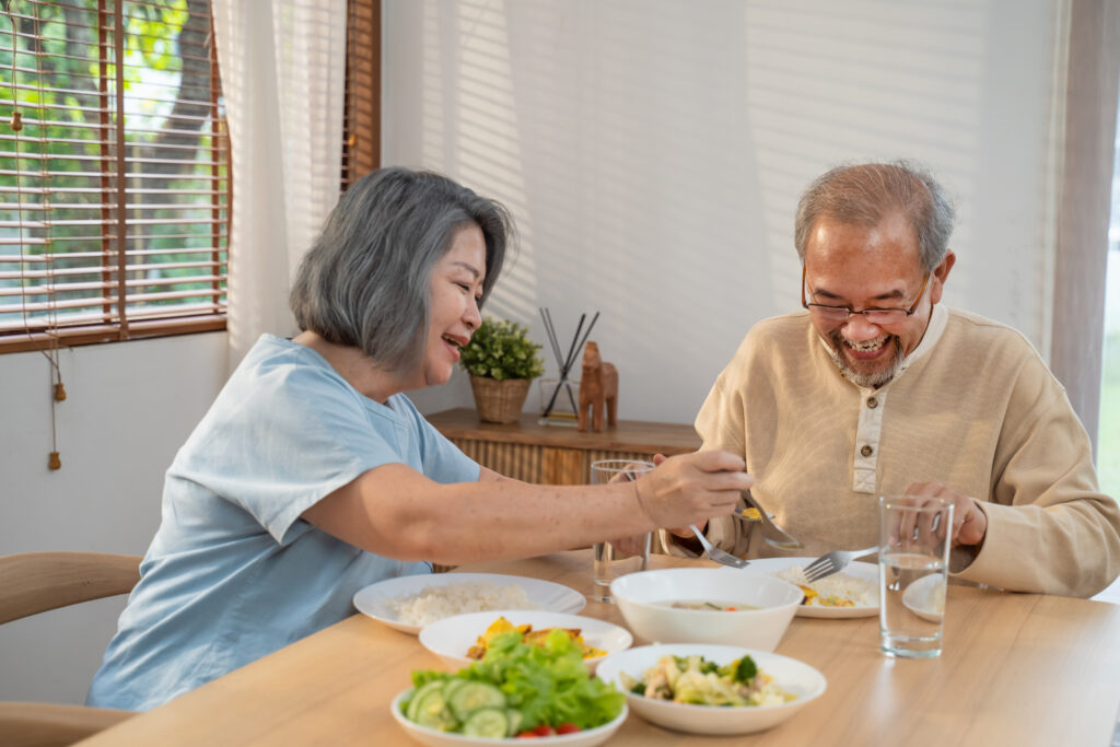 An older asian woman and older asian man sit at a dining table at home, putting food on their plates from several small bowls of food on the table.