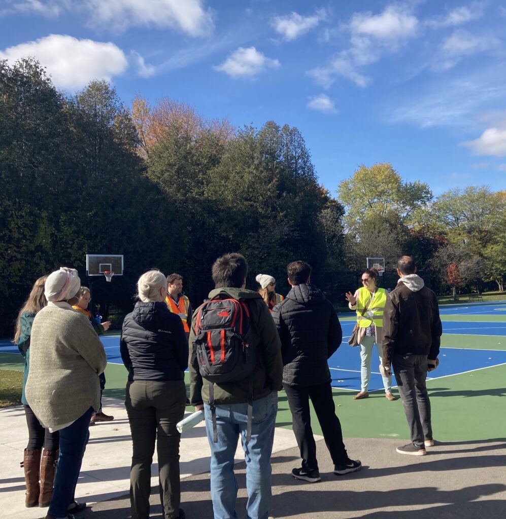 Woman in neon yellow safety vest speaks to seven adults in light jackets and two adults orange safety vests standing in a loose circle outdoors on a basketball playing court.