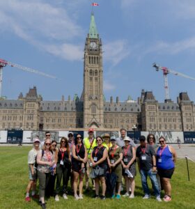 Group of 16 people in summer walking clothes, two wearing safety reflective vests, posing in front of the Peace Tower in Ottawa