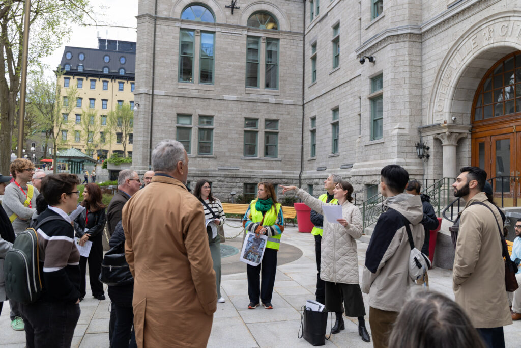 A group of people stand on the plaza in front of Quebec City Hall. A young dark-haired woman in a cream-coloured coat, black pants and black boots holds a paper in her left hand and is pointing away from the building: the group all is focused on looking where she is pointing.