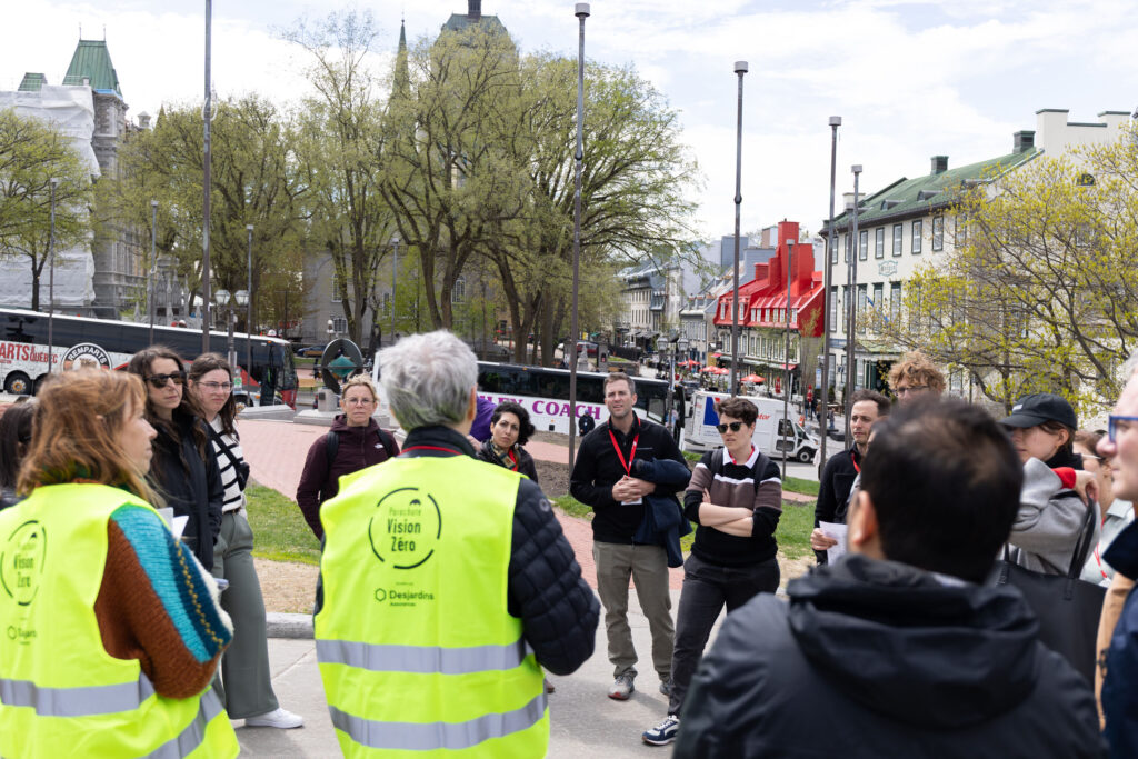 A man and a woman in the foreground with their backs to the camera, each wearing a reflective green safety vest with a Parachute Vision Zéro logo on back, are part of a circle of people standing on a plaza overlooking an old shopping street in Old Quebec City.